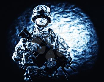 United States Marine Corps soldier in camouflage protective ammunition with hiding identity, armed with machine gun blue tone studio shot. Army infantry posing with weapon, spotlight in background