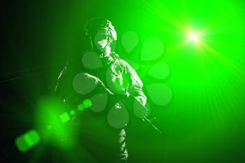 US marine raider in combat uniform with hidden face, armed with assault carbine low key, high contract studio shot on black background in blinding light. Equipped army soldier standing in darkness with weapon in hands