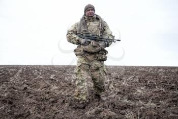 Army special forces machine gunner, elite commando in camouflage uniform, load carrier, standing on plowed field, holding medium machine gun, patrolling border line, observing area