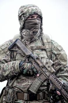 Irregular military mercenary, commando saboteur in camo jacket, wearing hood on head, armed service rifle, carrying tactical backpack during march, looking on distance