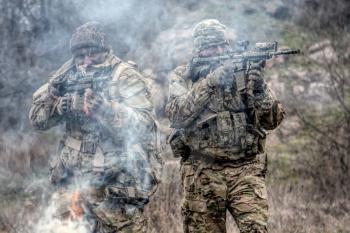 Two soldiers in camouflage uniform, wearing military ammunition, aiming service rifles, covering each other, shooting in competitors, attacking enemies trough smoke screen