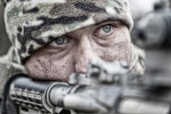 Close-up portrait of army infantryman, modern warfare combatant, young soldier with dirty, unshaven face, in camo bennie hat, aiming service rifle, controlling area with gunfire, looking in camera