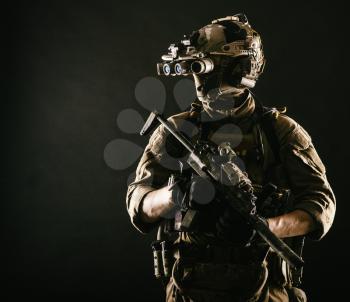 Army elite soldier with hidden behind mask and glasses face, in full tactical ammunition, looking aside, equipped night vision device, radio headset, armed short barrel rifle studio contour shot
