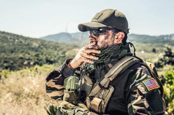 Army special forces soldier, SEALs fighter in camo uniform, shemagh scarf, ballistic glasses and cap, wearing tactical plate carrier, smoking cigarette, resting after fight in mountain area