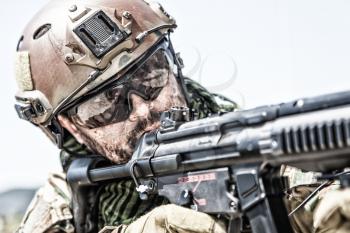 Close up portrait of army special forces soldier, commando rifleman, SEALs fighter with dirty face, wearing shemagh scarf, combat helmet and ballistic glasses, aiming and shooting with submachine gun