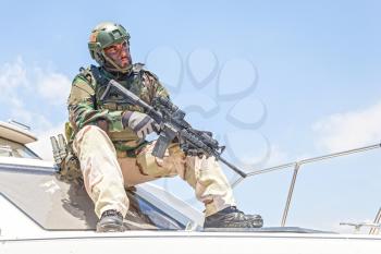 Navy SEALs fighter in battle uniform and helmet, holding service rifle in hand, looking into distance while sitting, resting or observing area from bow of speed boat during patrolling