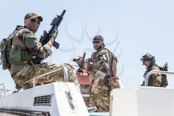 Army special operations soldiers, SEALs team, elite commando fighters group loaded with ammunition, wearing helmets and radio headset, armed assault rifles, standing together on speed boat stern