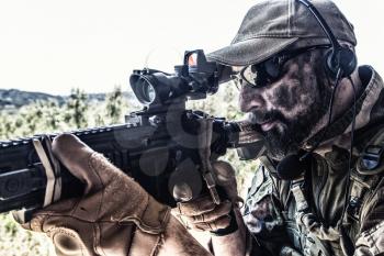 Army special forces soldier, modern combatant with dirty, unshaven face, in combat uniform, glasses and cap, equipped radio headset, aiming service rifle or light machine gun with optical sight