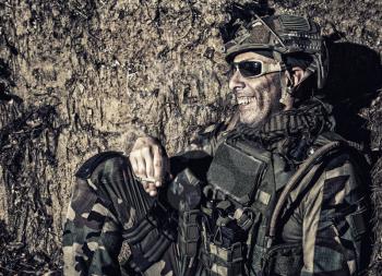 Smiling US army soldier, SEALs fighter, modern combatant in combat uniform, plate carrier, ballistic glasses and battle helmet resting after fight, sitting in trench at night and smoking cigarette