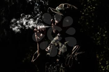 Commando soldier in camouflage uniform, ballistic glasses and bonnie, inhale cigarette smoke, smoking tobacco or marijuana joint in forest or jungles at night. Army infantryman relaxing after fight