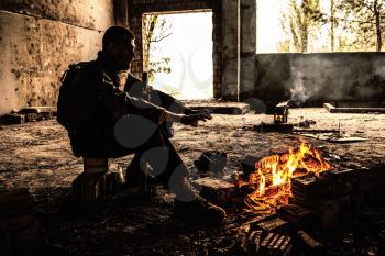 Special forces soldier after the fight sitting by the fire in ruined building warming his hands