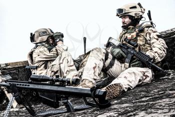 U.S. Navy SEAL infantrymen, commando marksman in battle uniform, armed with assault service rifle with optics sight, observing territory while sitting on observation post on roof of ruined building