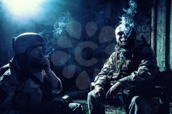 Tired after hard battle army soldiers, exhausted with fight Navy Seal rifleman sitting with assault rifle on knees, resting and smoking cigarette in abandoned building, low key, high contrast shoot