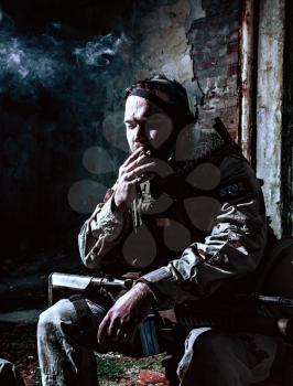 Tired after hard battle army soldier, exhausted with fight Navy Seal rifleman sitting with assault rifle on knees, resting and smoking cigarette in abandoned building, low key, high contrast shoot
