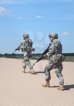 Pair of United states airborne infantry men with weapons moving patrolling desert. Sand and blue sky on background, sunlight, back view