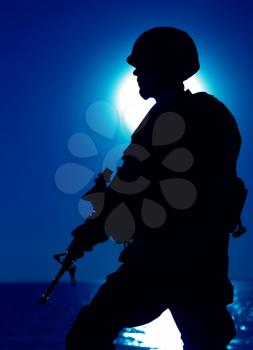 Silhouette of army infantry shooter patrolling seacoast with service rifle at night. Army special forces soldier, Marines fighter in helmet and uniform on night mission on seashore, sentry in darkness