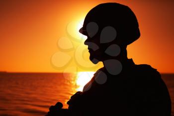Silhouette of army infantry soldier, special forces rifleman armed with service rifle, patrolling coastline at sunset. Coast guard fighter in combat helmet and tactical ammunition standing on seashore