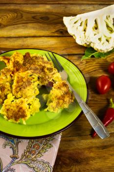 Baked cauliflower in breadcrumbs with cheese on a green plate, tomatoes, hot peppers, fork, napkin, wooden background, vertical