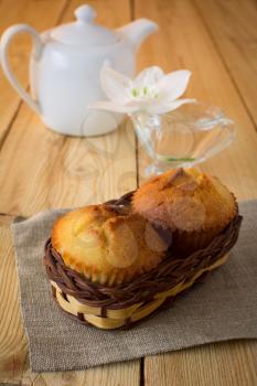 Two muffins in a small wicker basket on burlap, white flower and porcelain teapot on a wooden table, selective focus