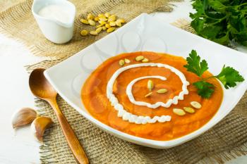 Roasted pumpkin squash vegetable soup with cream, pumpkin seeds and parsley in a white square plate on the burlap