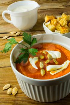 Pumpkin soup with cream and pumpkin seeds in the white bowl, cream in a creamer and croutons in a serving dish on the dark wooden background, close up, vertical