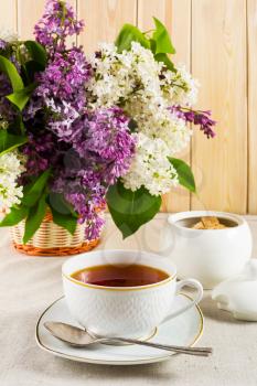 Cup of tea and branch of lilac flower in wicker basket on linen tablecloth. Spring tea time concept. Breakfast tea cup.