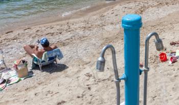 Man sitting on the portable chair. Tourist facing the shoreline reading a book. Picnic basket and mat lying on the ground. A shower post install near the sea. Relaxing beach resort destination