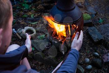 The man heats his hands in front of an open fire. Camping concept with outdoor open fire flames. Preparing barbecue fire. Camping fire and man in dark twilight moody photo. 