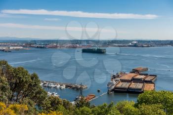 A container ship enter the Port of Tacoma. Barges can be seen in the forground and the Tacoma Dome in the distance.