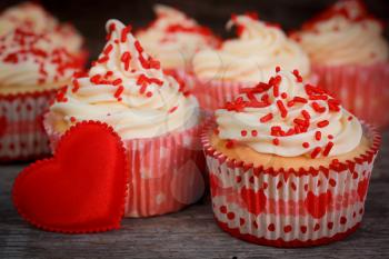 White cupcakes with vanilla icing and red candies with a red satin heart on a wooden background