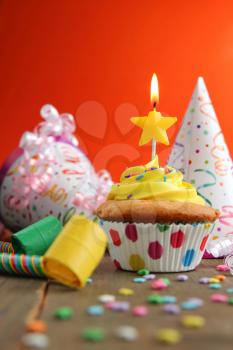 Vanilla cupcake with yellow frosting and a birthday candle with hats and blower on a wooden table and a orange background