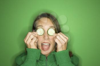 Portrait of young adult Caucasian woman on green background holding cucumber slices over her eyes.