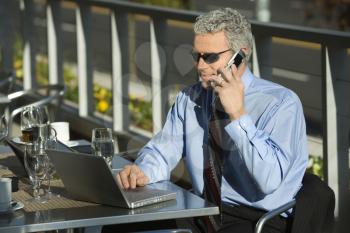 Royalty Free Photo of a Businessman Sitting at a Patio Table Outside With Laptop and Talking on His Cellphone