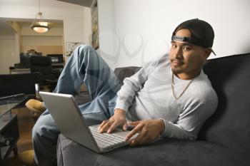 Royalty Free Photo of a Man Sitting on a Sofa Wearing a Cap Backwards and Typing on a Laptop