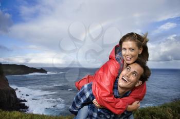 Caucasian mid-adult couple piggybacking by ocean with rainbow in background in Maui, Hawaii.