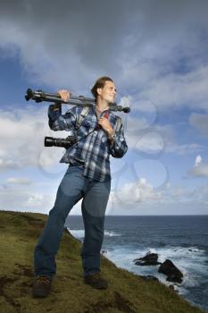 Caucasian mid-adult man standing with camera and tripod over his shoulder on cliff overlooking ocean in Maui, Hawaii.