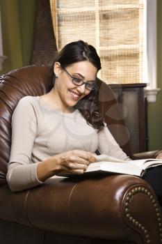 Caucasian/Hispanic young woman sitting in leather chair smiling and reading a book.