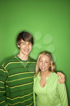 Portrait of Caucasian teen boy and girl standing against green background smiling with arms around eachother. 