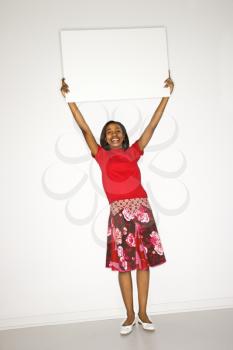 Portrait of African-American teen girl holding blank white sign above her head in front of white background.
