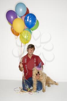 Royalty Free Photo of Man Sitting With a Dog Wearing a Party Hat and Holding Balloons