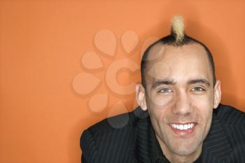 Head and shoulder portrait of Caucasian man in suit with mohawk smiling against orange background.