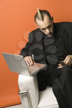 Royalty Free Photo of a Man in Suit with a Mohawk Typing on Laptop and Looking at His Cellphone While Sitting in a Chair