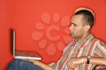 Caucasian man with mohawk holding laptop against red background.