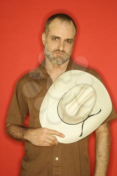 Caucasian man with beard and receding hairline holding cowboy hat to his chest against orange background.