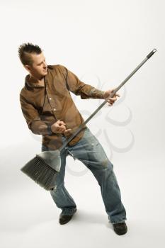 Caucasian man with mohawk playing broom like guitar against white background.