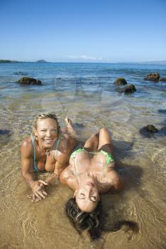 Pretty Caucasian mid adult women bodybuilders in bikinis sitting in shallow water on Maui beach.