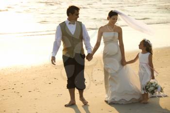Caucasian mid-adult bride, mid-adult groom and flower girl holding hands walking barefoot on beach.