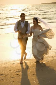 Royalty Free Photo of a bride and mid-adult groom holding hands running barefoot on beach.