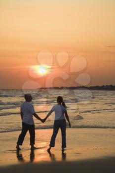 Mid-adult couple holding hands and walking on beach at sunset.