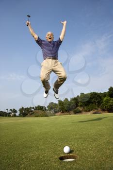 Caucasion mid-adult man holding golf club jumping in air cheering with golfball and hole in foreground.